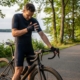 Rennradfahrer mit verspanntem Nacken steht neben seinem Rad mit weit gestreckter Lenkerposition am Wasser